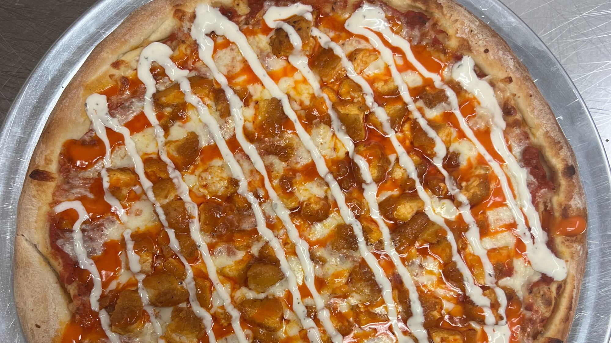 A buffalo chicken pizza on a steel pan, striped with hot sauce and ranch.