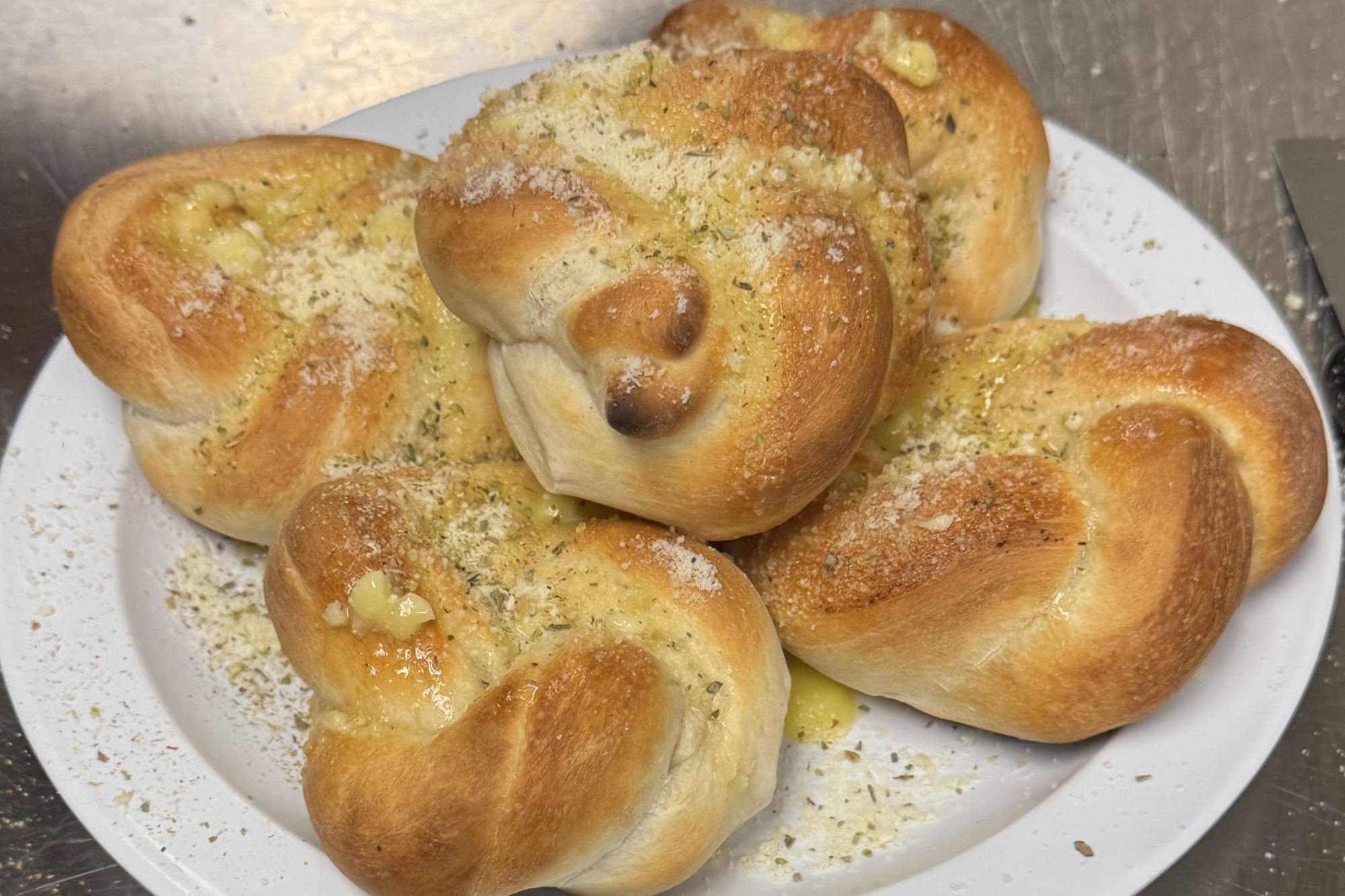 A plate of hand-tied garlic knots, gold from the oven, brushed with garlic butter and dusted with herbs and parmesan.