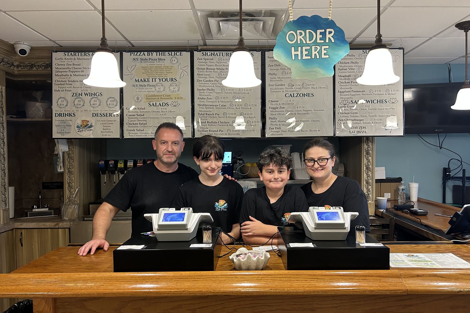 Akim and Hope Zendeli and their two children behind the counter at Zen Pizza, under the menu boards.